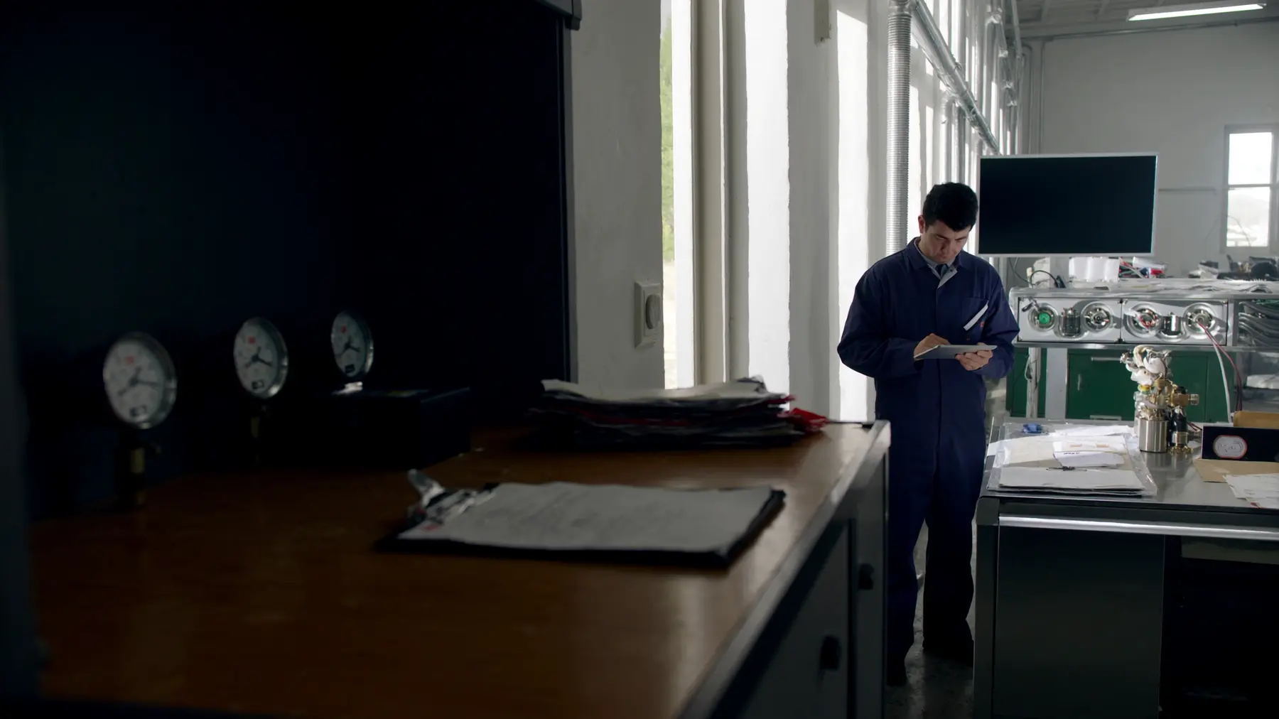 Werkstatt in einem Industriebetrieb: links im Schatten ein Stapel abgegriffener Ordner auf einem alten Stahlschrank mit runden Manometern, rechts im Tageslicht ein Instandhalter mit einem Tablet an einem Edelstahl-Arbeitstisch.